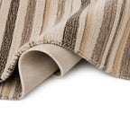 A close-up of a striped area rug with a textured, looped pile. The rug features vertical stripes in beige, tan, brown, and cream, with one corner rolled up to show the underside and backing.